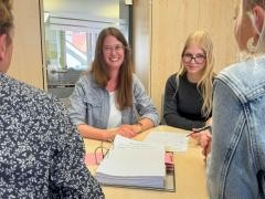Vier Personen sitzen an einem Tisch. Auf dem Tisch liegt ein Ordner. Zwei Personen sind nur von hinten zu erkennen. Zwei Personen sind ganz zu erkennen. Sie lächeln in die Kamera.