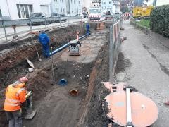 Straßenbauarbeiten mit offenem Graben für die Verlegung von Wasser- oder Abwasserleitungen. Mehrere Bauarbeiter sind im Einsatz, einer verdichtet mit einer Rüttelplatte den Boden. Ein blauer Rohrabschnitt und orangefarbene Anschlussstücke sind sichtbar. Die Baustelle ist mit Absperrgittern gesichert, im Hintergrund sind Wohnhäuser und Baumaschinen zu sehen.