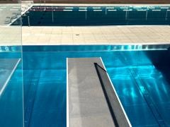 Ein Sprungbrett ragt zentral im Vordergrund über ein klar leuchtendes Schwimmbecken, dessen tiefblaues Wasser durch das Sonnenlicht schimmert. Der Schatten eines Geländers fällt diagonal über das Sprungbrett. Im Hintergrund trennt ein heller Fliesenstreifen das Becken vom dahinterliegenden Sport-Schwimmbecken mit mehreren markierten Bahnen. Die gesamte Szene wirkt ruhig, sonnig und geordnet – typisch für ein modernes Freibad oder ein Schwimmstadion.