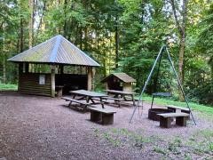 Eine Hütte im Wald. Davor stehen Bänke und ein Schwenkgrill.