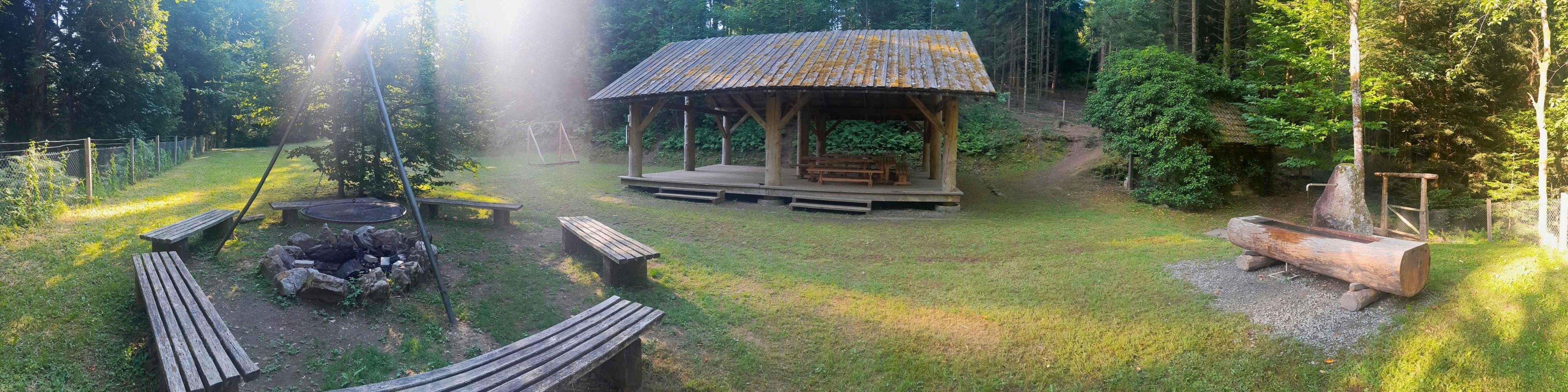 Links ist eine Grillstelle, in der Mitte eine Hütte, rechts ein Brunnen. Der Grillplatz ist von Wald umgeben.
