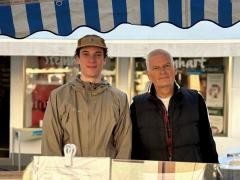 Zwei Männer stehen hinter einem Marktstand und lächeln in die Kamera.