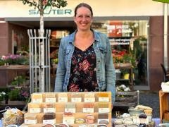 Eine Frau in Jeansjacke steht lächelnd hinter einem Marktstand. Vor ihr steht ein Aufsteller mit Seife.