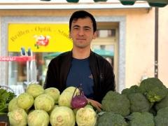 Ein Mann steht hinter einem Marktstand und lächelt in die Kamera. Vor ihm liegt verschiedenes Gemüse. Unter anderem Kohlrabi und Brokkoli.