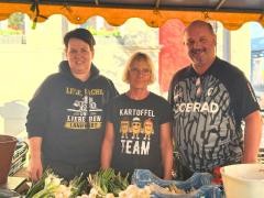 Drei Personen stehen hinter einem Marktstand und lächeln in die Kamera.