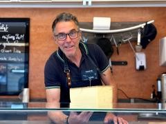 Ein Mann steht lächelnd hinter einem Marktstand. In der Hand hält er einen Käse.