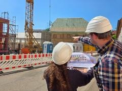 Zwei Personen mit Bauhelm stehen vor einem Gebäude, das sich gerade im Bau befindet. In der Hand halten sie einen Plan.