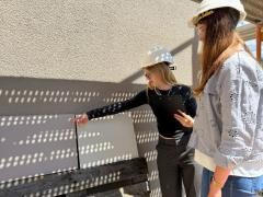 Zwei Personen mit Baustellen-Helmen stehen vor einem Haus. An der Hauswand lehnen zwei verschiedene Fließen. Eine Person  zeigt auf eine der Fließen.
