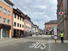 Die Fotografie zeigt eine belebte Innenstadtstraße in Waldkirch, Baden-Württemberg. Auf der rechten Seite befindet sich eine Bushaltestelle mit einem Haltestellenschild, das mit „Waldkirch Stadtmitte“ beschriftet ist. Daneben steht ein Mann mit Warnweste, der offenbar im öffentlichen Dienst arbeitet. Auf der Straße ist groß das Wort „BUS“ aufgemalt. Mehrere Menschen überqueren an einem Zebrastreifen die Straße. Links säumen bunte Gebäude mit Geschäften und Cafés die Straße. Einige Tische mit Sonnenschirmen stehen vor einem Lokal. Im Hintergrund sind weitere Häuser und ein leicht bewaldeter Hügel sichtbar. Der Himmel ist bewölkt mit leichten Auflockerungen.