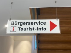 Auf einem weißen Schild steht "Bürgerservice" und "Tourist-Info". Ein roter Pfeil zeigt nach rechts. Das Schild hängt von einer Decke. Im Hintergrund ist eine Holztreppe zu sehen.