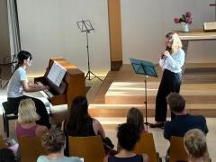 Am Klavier spielt eine Schülerin. Daneben steht eine Schülerin mit Blockflöte. Vor den beiden Musikerinnen sitzen Menschen.