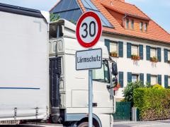 Ein LKW fährt an einem Haus vorbei. Vor dem Haus steht ein Tempo-30-Schild. Darunter steht "Lärmschutz".